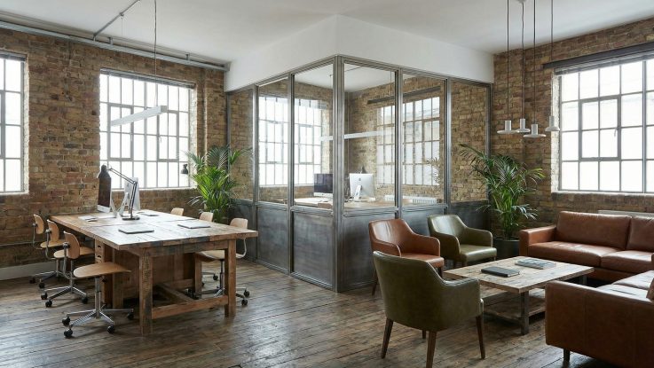 Ateş tuğlası duvarlı ofis alanında endüstriyel demir doğrama cam bölmeler ve doğal ahşap toplantı masası.