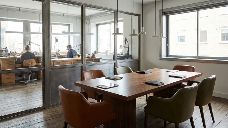 Masif ceviz toplantı masası, deri sandalyeler ve atölye üretim alanını gören şeffaf cam bölme sistemleri.