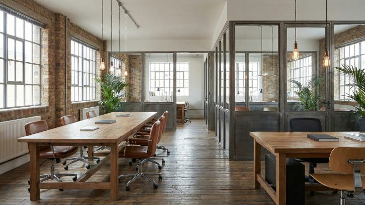 Masif ahşap masa üzerine sarkan filament ampullü rustik aydınlatmalar ve endüstriyel cam ofis bölmeleri.